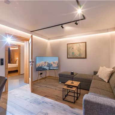 A modern living room with a gray couch and a television. The space is bright and friendly, with warm lighting and wooden details.