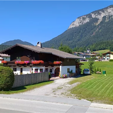 Ein charmantes bayerisches Haus mit Balkonblumen liegt idyllisch in der Natur. Im Hintergrund sind sanfte Hügel und eine klare blaue Luft zu sehen.