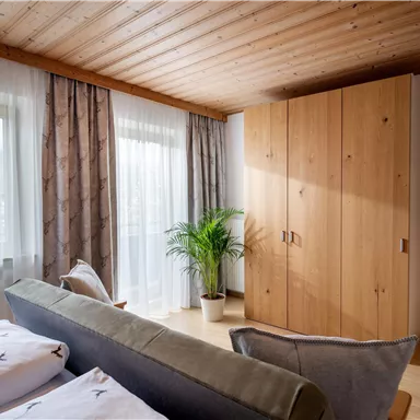 Ein gemütliches Schlafzimmer mit Holzdecke und hellen Vorhängen. Der Raum ist mit einem großen Kleiderschrank und einer Pflanze dekoriert.
