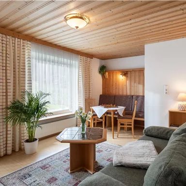 Ein gemütliches Wohnzimmer mit einem Sofa, einem Tisch und Stühlen. Große Fenster lassen viel Licht herein und verleihen dem Raum eine einladende Atmosphäre.