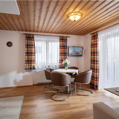 Ein gemütlicher Wohnraum mit Holzdecke, großer Fensterfront und bunten Vorhängen. Ein Esstisch mit modernen Stühlen und eine kleine Küchenecke sind ebenfalls sichtbar.