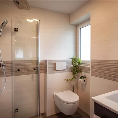 Ein modernes Badezimmer mit einer Dusche, einem weißen Waschbecken und einer Toilette. Große Fenster sorgen für viel Tageslicht und eine grüne Pflanze verleiht eine frische Note.