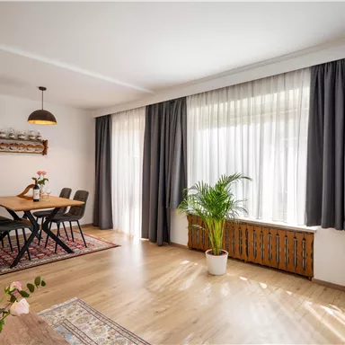 Ein helles Wohnzimmer mit einem Esstisch, Stühlen und einer großen Fensterfront. Pflanzen und ein Fernseher verleihen dem Raum eine gemütliche Atmosphäre.