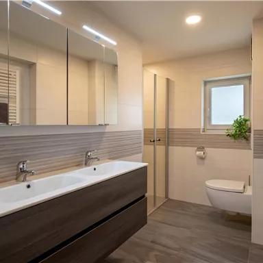 Ein modernes Badezimmer mit zwei Waschbecken und großflächigen Spiegeln. Es hat helle Fliesen und eine Dusche mit Glasabtrennung.