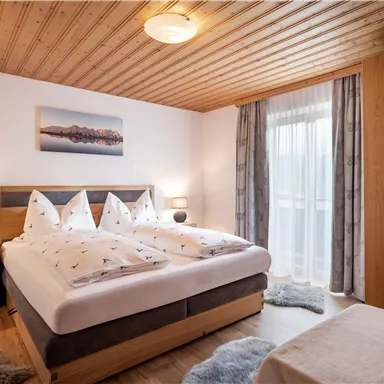 Ein gemütliches Schlafzimmer mit einem großen Bett und dekorativen Kissen. Die Holzdetails und das Fenster sorgen für eine warme Atmosphäre.
