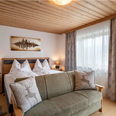 Ein gemütliches Schlafzimmer mit einem Doppelbett und zwei Nachttischen. Große Fenster und ein Sofa sorgen für eine einladende Atmosphäre.