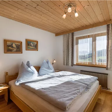 Ein gemütliches Schlafzimmer mit Holzdecke und großen Fenstern. Es gibt ein Doppelbett, zwei Kissen und dekorative Bilder an der Wand.