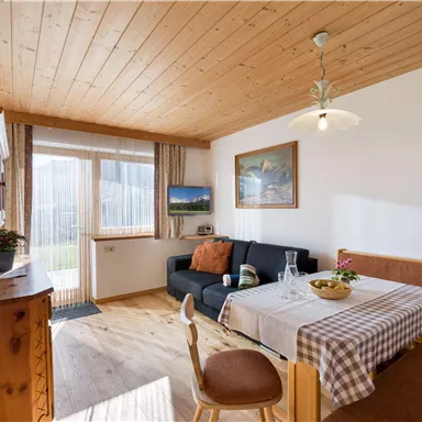 Ein gemütlicher Wohnraum mit Holzboden und Holzdecke. Es gibt ein Essplatz mit einem Tisch und einer Couch sowie große Fenster, die viel Licht hereinlassen.
