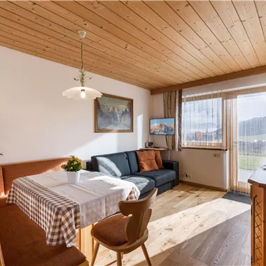 Ein gemütliches Wohnzimmer mit einem Essbereich, einer dunkelblauen Couch und großen Fenstern. Der Holzboden und die Holzdecke verleihen dem Raum eine warme Atmosphäre.