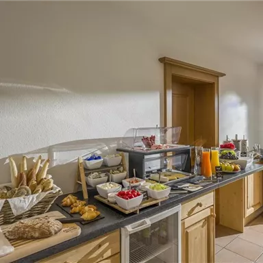 An inviting breakfast bar with fresh rolls, cold cuts, and fruit. The room has large windows that offer a view of the surrounding nature.