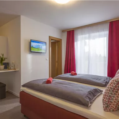 A cozy room with two beds, a television, and large windows. Red curtains and a small kitchenette give the space an inviting atmosphere.