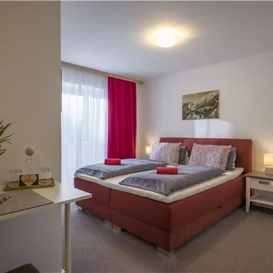A cozy bedroom with two beds, decorated with red pillows. The walls are light, and large windows provide plenty of daylight.
