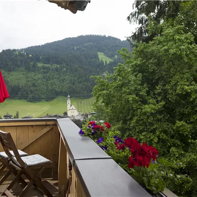 A balcony with a red parasol and blooming flowers. In the background, green hills and a church are visible.