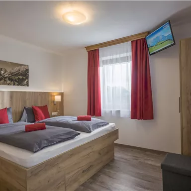 A modern bedroom with two assembled beds and red pillows. The room features wooden details, a window with curtains, and a television on the wall.