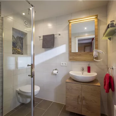 A modern bathroom with a shower, a sink, and a mirror. The walls are tiled in light colors, and there are decorative elements like a picture and towels.