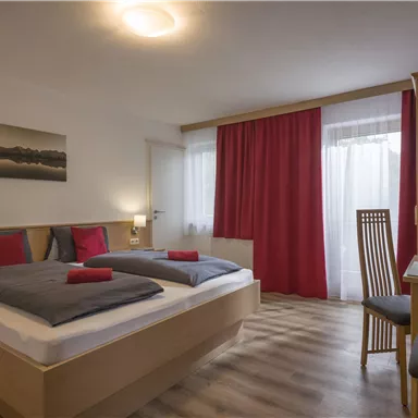 A cozy hotel room with two beds, modern furniture, and a desk. Large windows and red curtains create an inviting atmosphere.