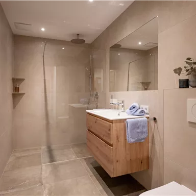 A modern bathroom with a wooden vanity and a large mirror. The walls are in neutral tones, and there is a shower with a glass sliding door.