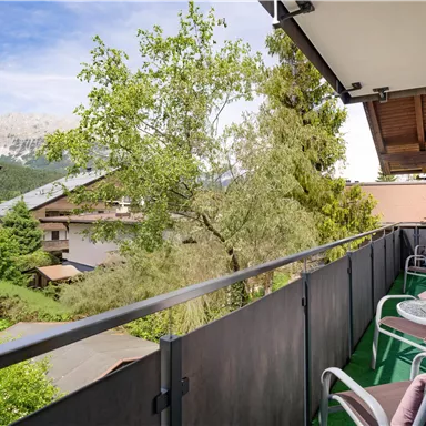 A balcony with a view of green trees and the mountains in the background. On the balcony, there are chairs and a table, ideal for relaxing.