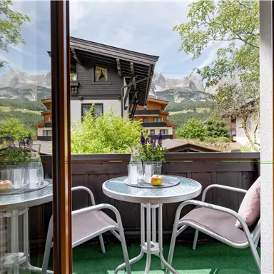A balcony with a small table and two chairs. In the background, trees and mountains can be seen.