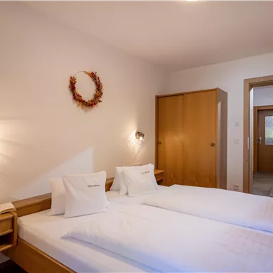 A bright bedroom with two single beds and modern wooden interior. The room feels cozy and inviting with warm light and a decorative wreath on the wall.