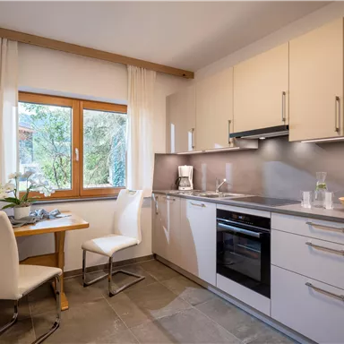 A modern kitchen with white furniture and a large window. A table and chairs are available, as well as appliances for kitchen use.