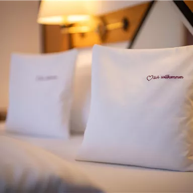 A cozy bedroom with two white pillows on the bed. The pillows are embroidered with the inscription "Welcome".