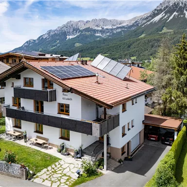 A modern house with solar panels and a beautiful garden. In the background, impressive mountains and green meadows are visible.