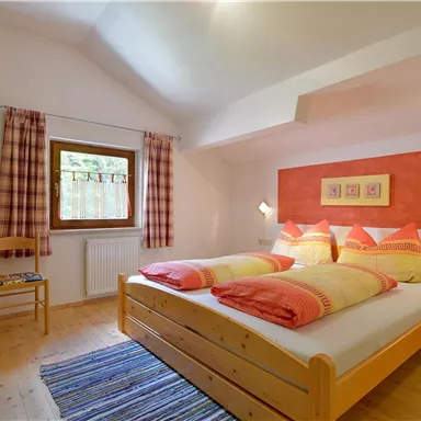 Ein gemütliches Schlafzimmer mit einem Doppelbett und leuchtend roten Kissen. Die Wände sind in sanften Farben gehalten, und ein Fenster sorgt für Tageslicht.
