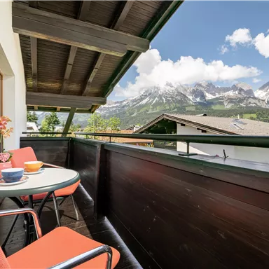 A balcony with a table and chairs, surrounded by mountains and beautiful scenery. The sky is blue and the clouds are lightly scattered.