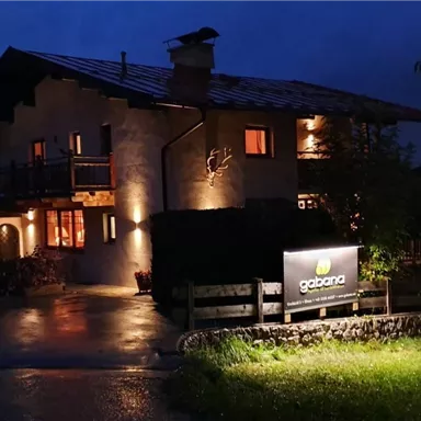 A cozy house at night, lit by warm lights. In the foreground, there is a sign with the inscription "gazana".