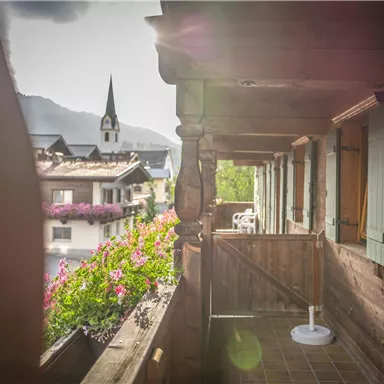 A balcony with wooden elements and blooming plants. In the background, there is a small church and the mountains.