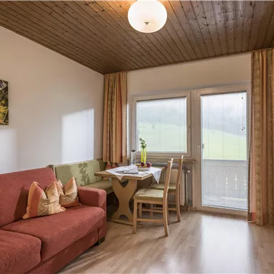 A cozy living space with a red sofa and a dining table. The large windows offer a view of the green landscape.