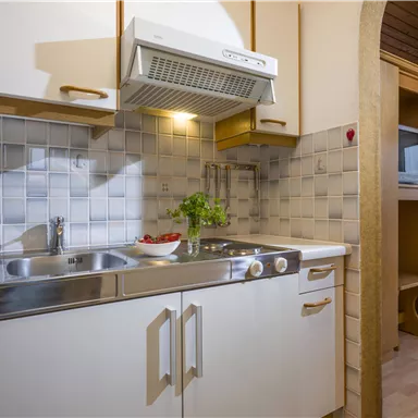 A modern kitchen corner with a sink, stove, and a microwave. The walls are tiled with light tiles and the lighting creates a friendly atmosphere.