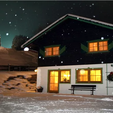 A pretty, dark blue house in the snow with illuminated windows. In the background, gentle, snow-covered hills and a lit tower can be seen.