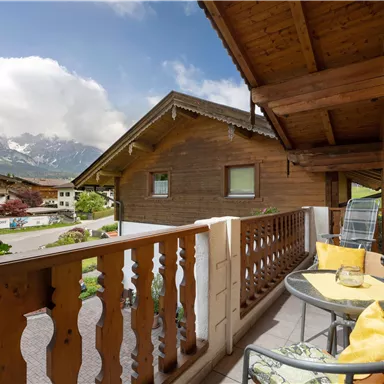 Ein gemütlicher Balkon mit einem Tisch und Stühlen, dekoriert mit gelben Kissen. Im Hintergrund sind die Holzhäuser und die beeindruckenden Berge zu sehen.