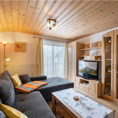 Ein gemütliches Wohnzimmer mit einer grauen Couch und einem Couchtisch. An der Wand befindet sich ein Fernseher und große Fenster lassen viel Licht herein.