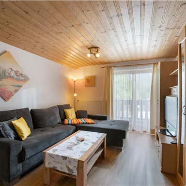 Ein helles Wohnzimmer mit einem gemütlichen Sofa und einem Couchtisch. Große Fenster lassen viel Licht herein und es gibt eine Fernsehecke sowie eine Holzdecke.