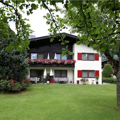 A charming house with red shutters and colorful flowers. The well-maintained garden is surrounded by trees.