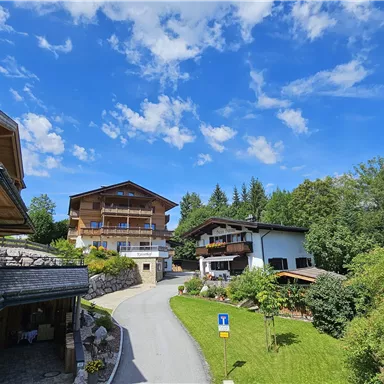 A picturesque landscape with several houses surrounded by green trees. The sky is blue with a few white clouds.