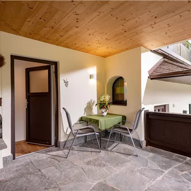 A cozy balcony with a table and two chairs. In the background, a fireplace and nature can be seen.