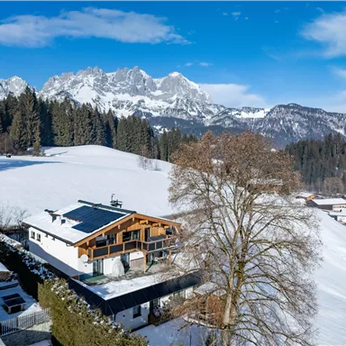 Eine malerische Winterlandschaft mit schneebedeckten Bergen und einem Holzhaus. Im Vordergrund stehen grüne Bäume und sanfte Hügel.