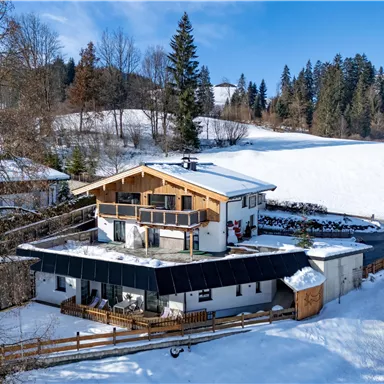 A snow-covered house in a quiet landscape. In the background, tall trees and gentle hills are visible.