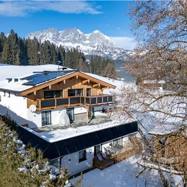 Ein charmantes Bauernhaus in den Alpen, umgeben von schneebedeckten Bergen. Der blaue Himmel und die sonnige Landschaft schaffen eine idyllische Winteratmosphäre.