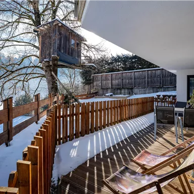 Eine gemütliche Terrasse mit Liegestühlen und einem Sichtschutz aus Holz. Im Hintergrund steht ein Baumhaus und der Boden ist mit Schnee bedeckt.