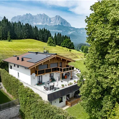 A modern house surrounded by lush greenery and trees. In the background, majestic mountains rise under a clear sky.
