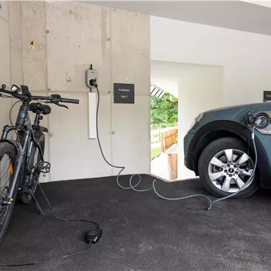 A modern garage environment with an e-bike and an electric car, both of which are charging. On the wall, there is a charging station for electric vehicles.