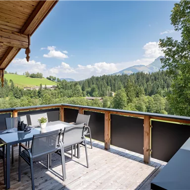 A cozy terrace with a table and chairs, surrounded by green forests and mountains. The sky is blue with white clouds.