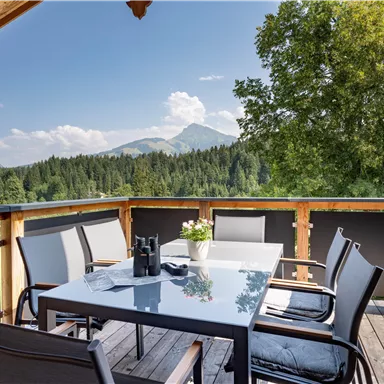 A cozy balcony with a table and chairs, surrounded by green mountains and trees. The sky is blue with some clouds.