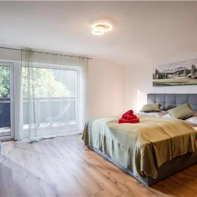A modern bedroom with a double bed and a cozy sofa. Large windows provide plenty of daylight and a view of the greenery.