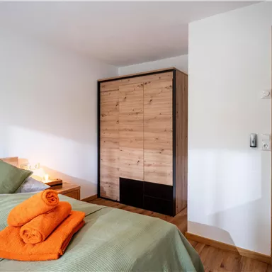 A modern bedroom with a cozy bed and fresh towels. The space is bright and welcoming, featuring a wooden closet and a view of the adjoining room.